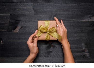 Female Hands Holding Craft Gift Box With Gold Ribbon In The Black Wooden Background