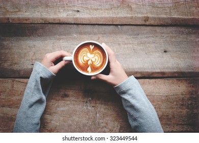 A Female Hands Holding Cup Of Coffee Latte Art On Wood Table. Above View And Vintage Effect.