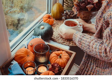 Female Hands Holding Cup Of Tea Near Autumn Cozy Mood Composition On The Windowsill. Pumpkins, Cones, Candles On Wooden Tray. Autumn, Fall, Hygge Home Interior Decor. Selective Focus. Copy Space