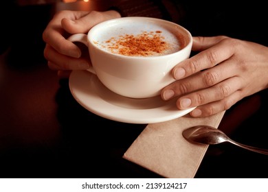 Female hands holding a cup of turkish traditional winter hot drink salep with cinnamon. White ceramic mug on plate and little spoon on napkin. Cozy atmosphere. Close-up.