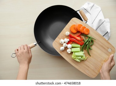 Female Hands Holding Cutting Board With Vegetables And Pan On Wooden Table, Top View