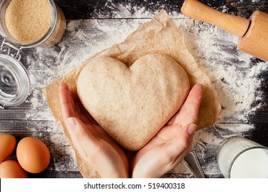 female hands holding dough in heart shape top view. Baking ingredients on the dark wooden table
