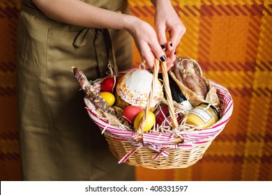 Female Hands Holding Easter Basket With Colorful Easter Eggs, Cake, Red Wine, Jamon Or Jerky And Dry Smoked Sausage. Food Gift Set For Celebrating Easter