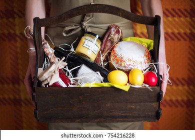 Female Hands Holding Easter Basket With Colorful Easter Eggs, Cake, Red Wine, Hamon Or Jerky And Dry Smoked Sausage. Food Gift Set For Celebrating Easter