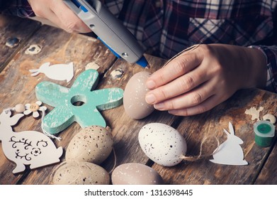 Female Hands Holding Easter Egg And Hot Glue Gun, DIY Composition On Rustic Wooden Table