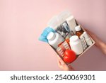 Female hands holding a first aid kit box with essential medical supplies on a pink background. Medical donations, first aid kit, home delivery of medications, online pharmacy concept.