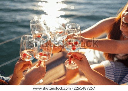Female hands holding glasses of champagne with raspberries on board the boat. Sunset sky and sea on the background. Making a celebratory toast with sparkling wine. Birthday, holiday, party and friends