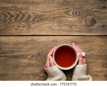 Female hands holding a mug of hot black tea. Cold winter white warm clothes. Top view. Aged rustic wooden background