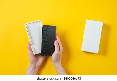 Female Hands Holding New Smartphone In The Box. Unboxing Top View On Yellow Background, Minimalism