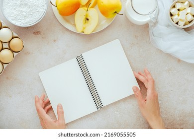 Female Hands Holding An Open Cook Book With Ingredients Flour, Milk, Eggs, Apples, Butter. Recipes For Autumn Vegan Baking. Flat Lay, Top View. High Quality Photo