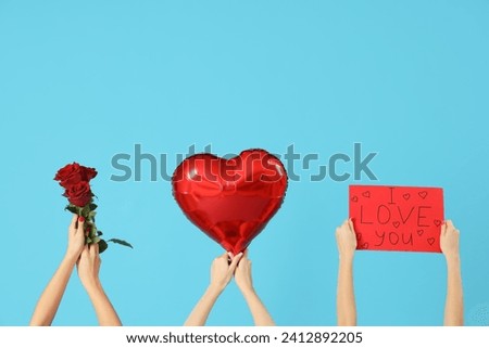 Female hands holding paper sign with text I LOVE YOU, heart-shaped balloons and roses on blue background. Valentine's Day celebration