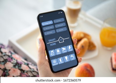 weibliche Hände, die ein Telefon mit App Smarthome auf dem Bildschirm über den Frühstückstisch halten 