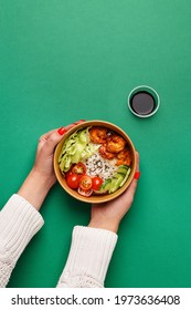 Female hands holding poke with shrimps, rice and vegetables in cardboard bowl over green background. Top view, flat lay, copy space. Traditional Hawaiian dish