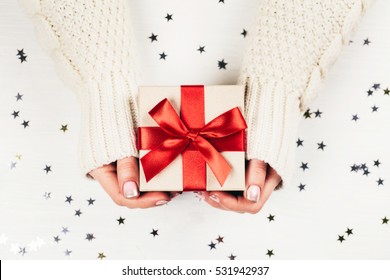 Female hands holding present with red bow on white rustic sparkling background. Festive backdrop for holidays: Birthday, Valentines day, Christmas, New Year. Flat lay