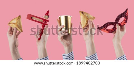 Female hands holding Purim celebration acessories and symbols. Jewish carnival background. Mask, traditional jewish hamantaschen cookies cakes, rattle noisemaker on pink banner