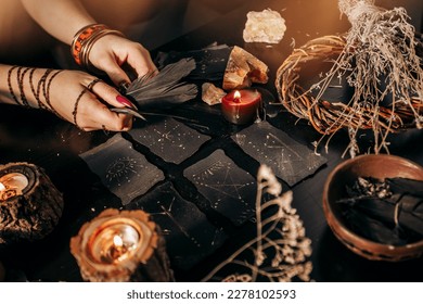 Female hands holding raven feathers against backdrop magical black patterned cards   lit candles in close  up  Halloween
