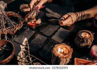 Female hands holding raven feathers against backdrop magical black patterned cards   lit candles in close  up  Halloween