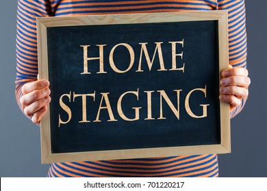 Female hands holding small black chalkboard in front of the body closeup with words Home Staging. On gray background with copy space