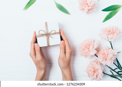 Female Hands Holding Small Gift Box Next To Bouquet And Green Leaves On White Wooden Table, Top View.