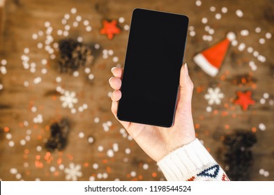 Female hands holding smart mobile phone with oled display on wooden background with Christmas gifts snowflakes and snow magic fairy tale light effect. Happy New Year and Xmas Flat lay