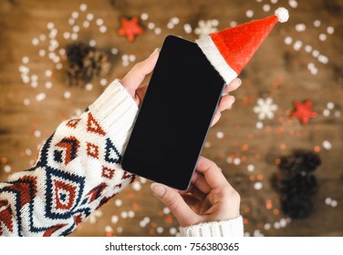 Female Hands Holding Smart Mobile Phone With Oled Display On Wooden Background With Christmas Gifts Snowflakes And Snow. Happy New Year, Hat Santa On Mobile Phone, Flat Lay Composition Top View.