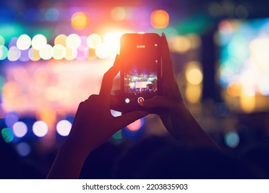 Female Hands Holding Smart Phone To Record Video During A Live Concert. Blurry Lights On The Background. Live Music, Crowd Of Fans With Cell Phone In Hands At Rock Concert In Spotlight Lighting.