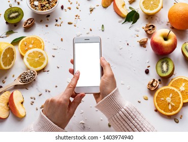 Female Hands Holding Smartphone On Healthy Food Background, Woman Using Phone Search Mobile Apps With Diet Nutrition Plan Cooking, Vegan Fruit Granola Seeds On White Table, Top View, Mock Up Screen