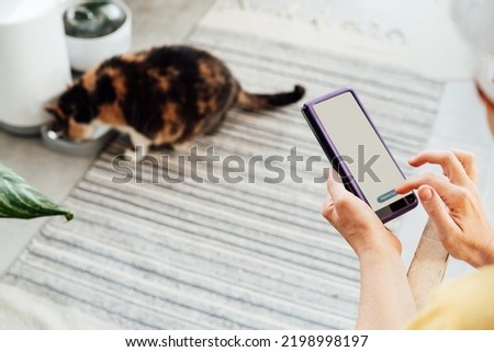 Female hands holding smartphone and using application to control cat food dispenser. Cat eating from smart feeder in cozy home interior on background. Home life with a pet. Healthy pet food diet.