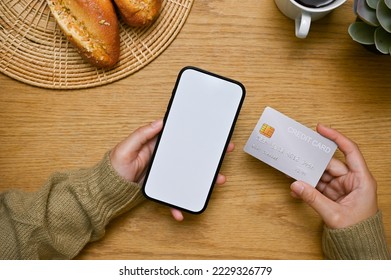 A female hands holding a smartphone white screen mockup and a credit card over wooden tabletop. top view