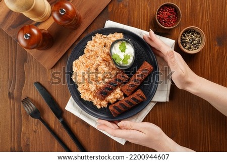 Female hands holding south european sausages cevapcici made of ground meat and spices on black plate on dark wooden board, vegan substitute made of soy protein, with rice and sour cream, top view