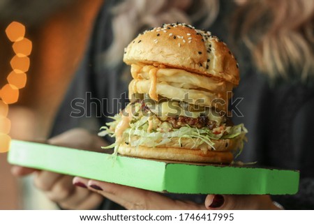Female hands holding spicy jalapeno burger. Woman in the black uniform serving homemade hanburger with pepper slices, beef patty, lettuce, fried onion rings on sesame buns on the cutting board. Image