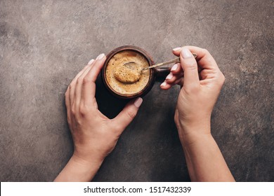 Female hands holding spoon and stirring hot coffee on a dark brown rustic background. Top view, flat lay