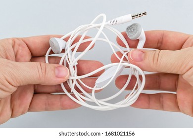 Female hands holding tangled earphones on a gray background. White tangled headphones. Modern personal device for music and communication. Front view. 