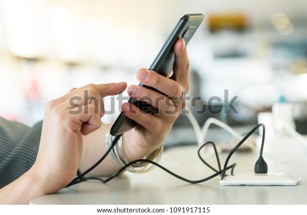 Female\
hands holding and using smartphone while charging it in a public\
place using electric plug and a charging cable\
