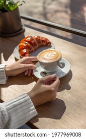 Female Hands Holding White Cup Of Cappuccino With A Croissant On A Wooden Table In Front Of A Window In A Coffee Shop. Morning Breakfast, Coffee Break. Espresso Bar. Tasty Beverage And Sweet Bakery