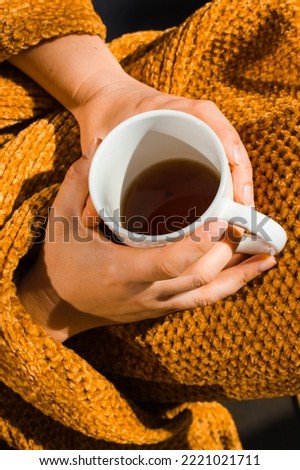Female hands holding white cup mug in hands top view. Woman wrapped in yellow plaid is drinking hot beverage, black tea in autumnal day. Good morning. Autumn, winter cozy sunny day. Relax leisure mood
