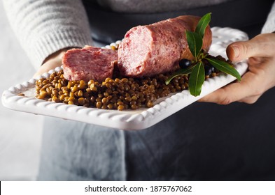 Female Hands Holding White Plate With Italian Large Pork Sausage Cotechino Served  With Lentils.