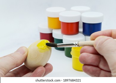 Female hands holding wooden Easter egg and paint it with yellow paint on background paint cans with different colors. Preparing decorations for Easter, creativity with children, traditional symbols
