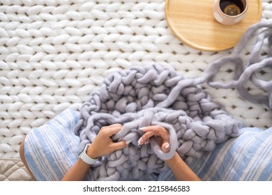 Female Hands Knitting Warm Plaid Merino Wool Ribbon Yarn Sitting On Bed With Hot Tea Top View Closeup. Art Woman Creating Texture Big Knit Blanket With Large Thread Comfortable Soft Woven Material