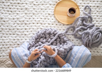 Female Hands Knitting Warm Plaid Merino Wool Ribbon Yarn Sitting On Bed With Hot Tea Top View Closeup. Art Woman Creating Texture Big Knit Blanket With Large Thread Comfortable Soft Woven Material