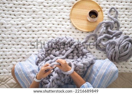 Female hands knitting warm plaid merino wool ribbon yarn sitting on bed with hot tea top view closeup. Art woman creating texture big knit blanket with large thread comfortable soft woven material