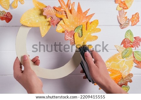 Female hands making decorative autumn paper wreath. Using stapler to fix leaves in circle on cardboard form. top view. DIY handmade autumn decorations