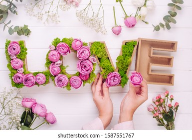 Female hands making volume letters LOVE with stabilized moss and pink roses on white wooden background. Top view, flat lay.