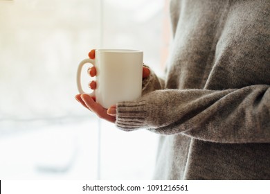 Female hands with a mug of beverage. Beautiful girl in grey sweater holding cup of tea or coffee. Empty mug for your design. - Shutterstock ID 1091216561
