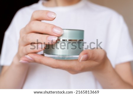 Female hands with natural pink manicure holding a transparent cream jar. Woman in white holds beauty product. Concept of body care and skin protection. Moisturizing lotion closeup. Facial skincare
