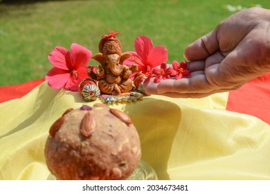 Female Hands Offering Flowers To Lord Ganesha At Home. With Selective Focus On Ganesha And Laddu Prashad In Foreground. Ganesh Chaturthi Pooja Celebrations At House