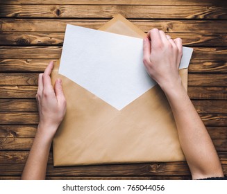 Female Hands Open A Large Brown Envelope And Take Out A Letter From It. Wooden Background.