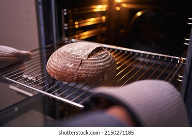 Female Hands In Oven Mitts Take Round Rye Bread Out Of The Oven