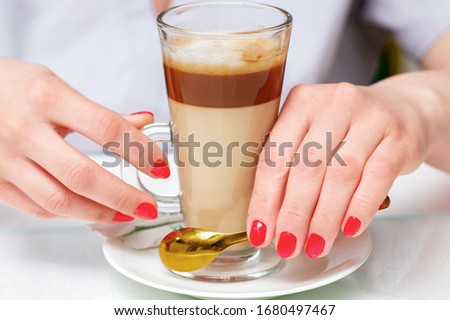 Female hands with perfect red manicure holds cup of coffee cappuccino close up.