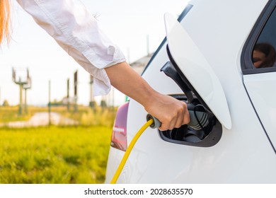 Female hands plugging in the charger into a socket of white electric car at a charging station near an electric power plant
